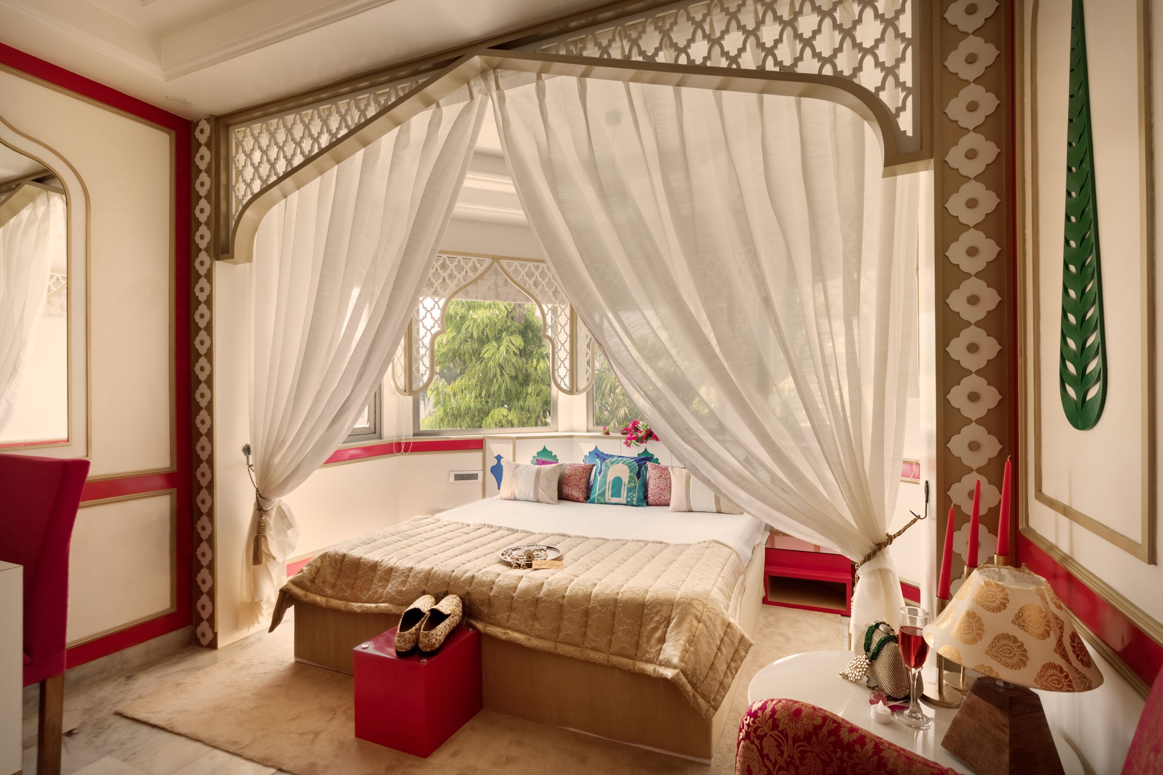 Une chambre à coucher avec un lit à baldaquin, un pouf rouge, une table de chevet avec une lampe et une fenêtre donnant sur un paysage verdoyant.
