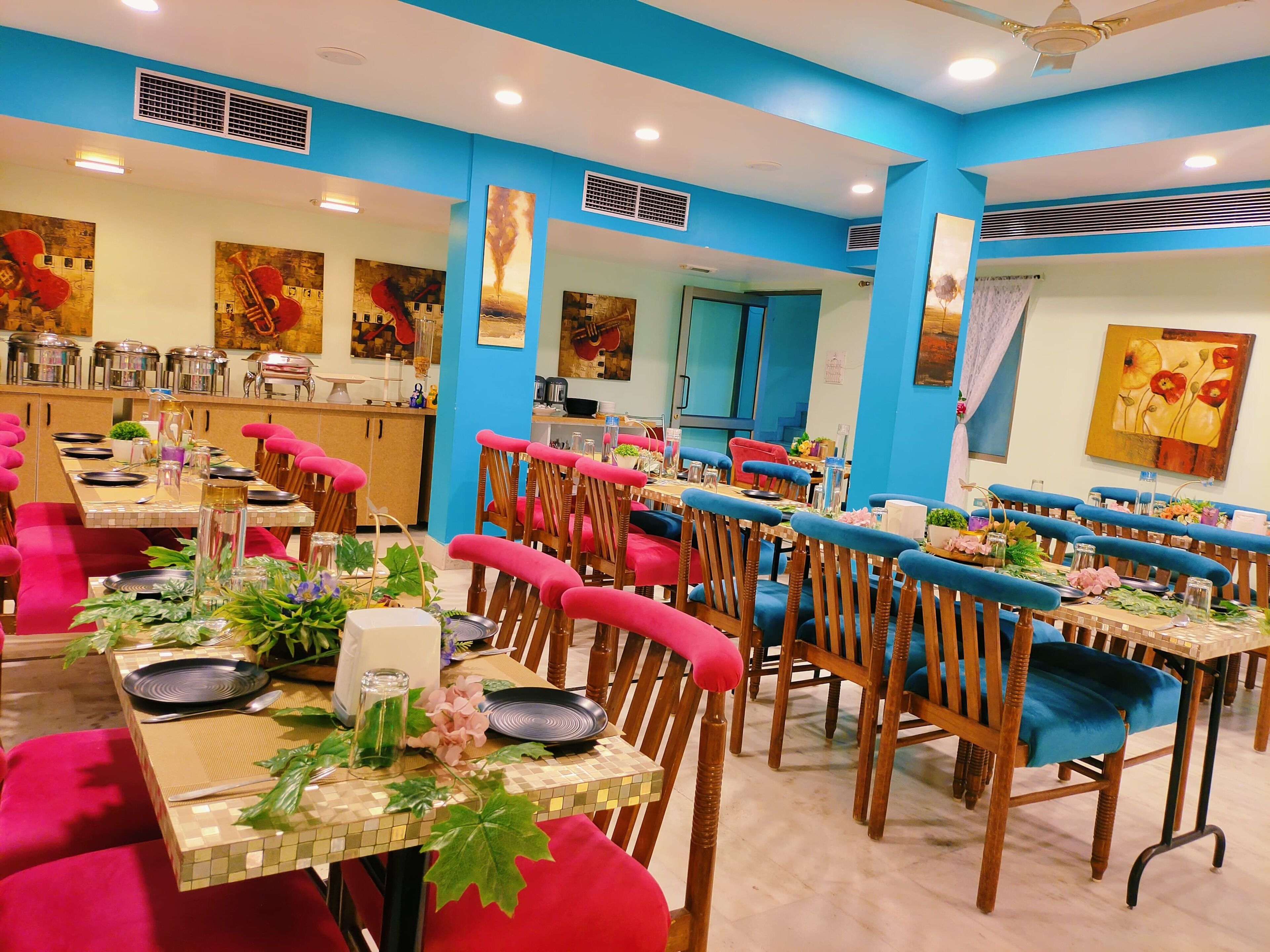 Un espace repas avec des tables disposées pour accueillir quatre personnes, agrémenté de chaises rembourrées aux couleurs rouge et bleu, et un coin cuisine équipé d’appareils électroménagers en acier inoxydable.