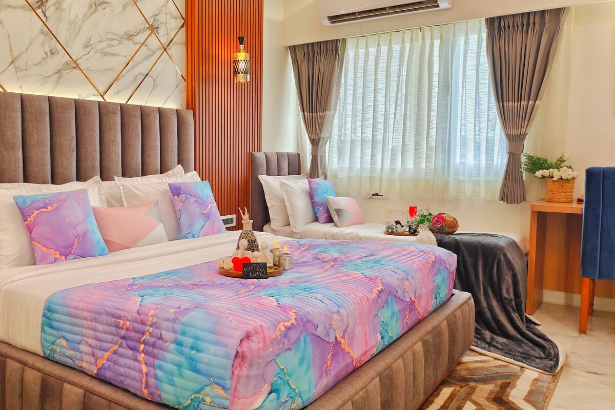 Une chambre d’hôtel moderne avec un grand lit, un plus petit lit, une tête de lit en bois, un panneau décoratif mural, une table de chevet avec un vase et une fenêtre avec des rideaux.