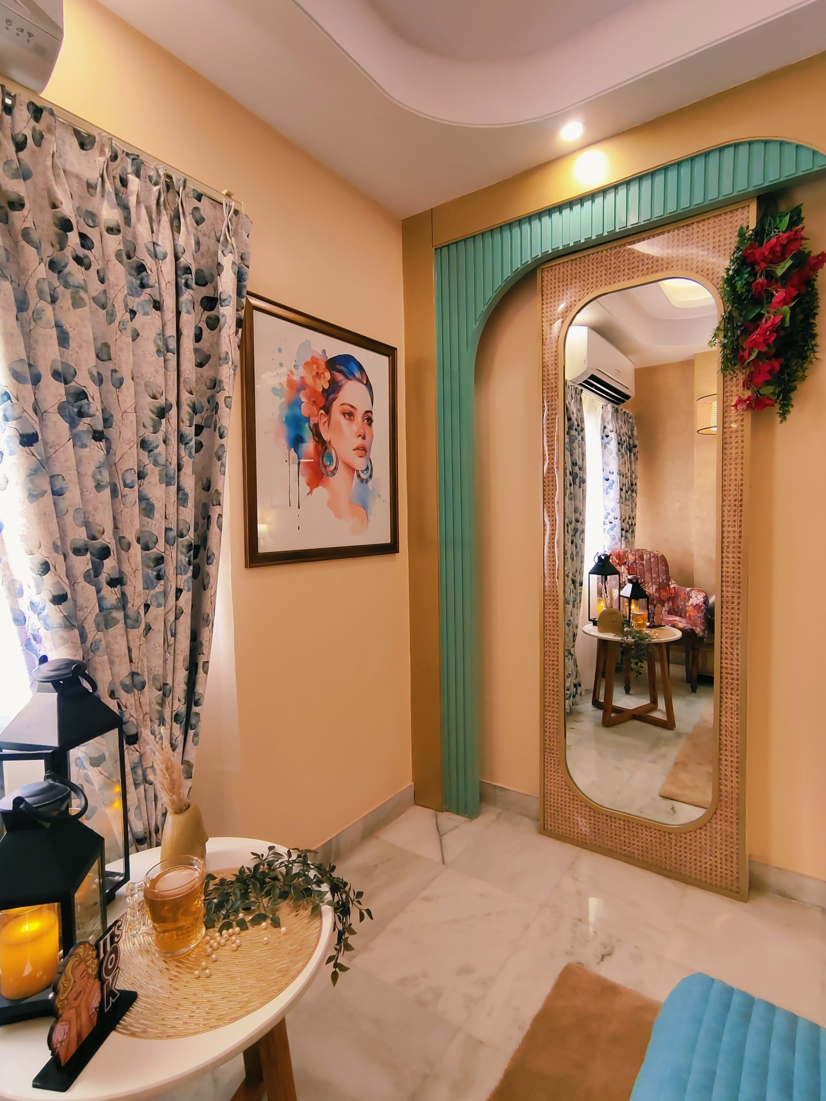 A room with a large mirror, a framed artwork, a table with a candle and a vase, and a blue chair.