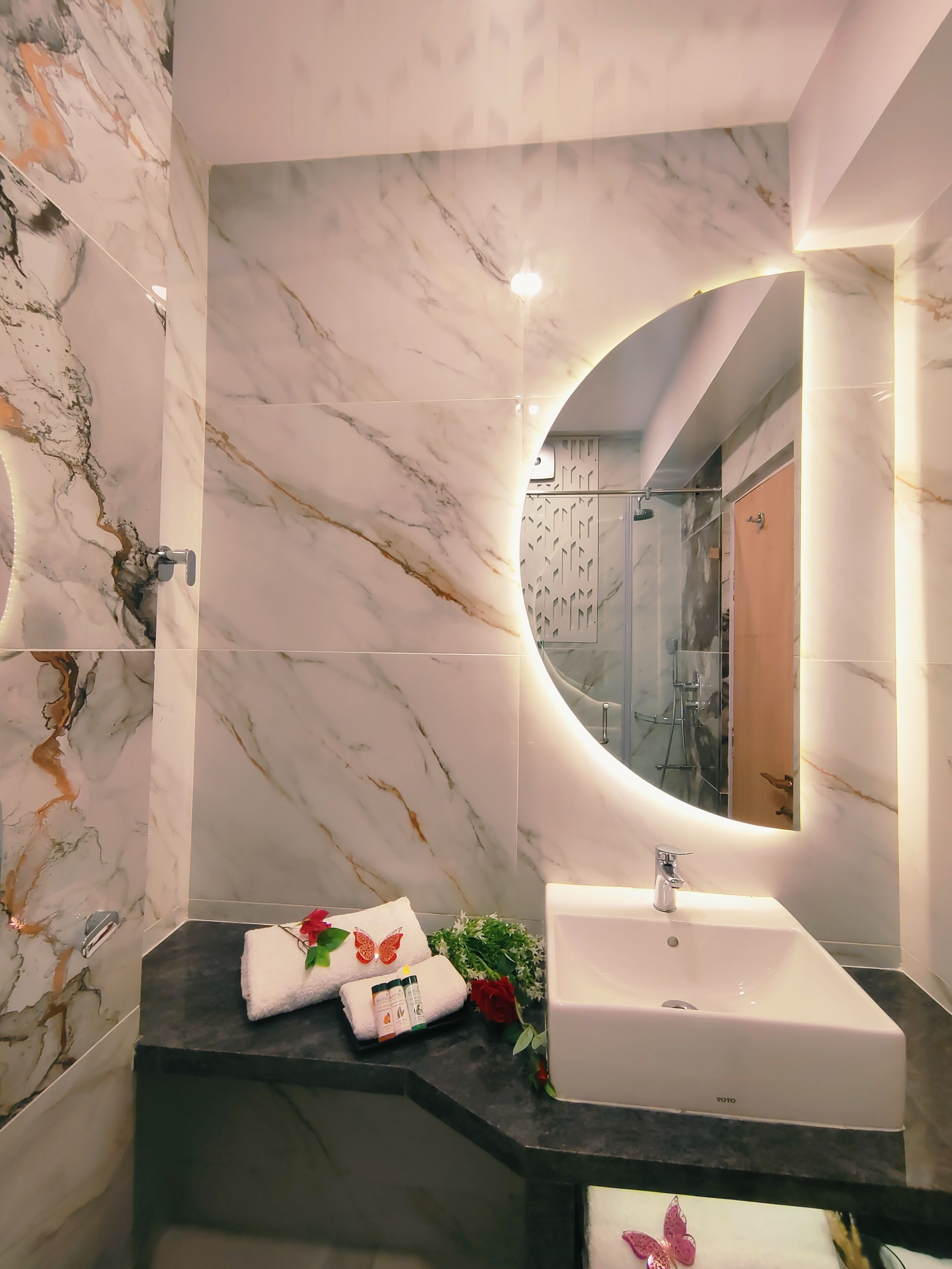 Une salle de bain moderne avec un mur en marbre, un miroir rond, un lavabo blanc et un plan de travail noir.