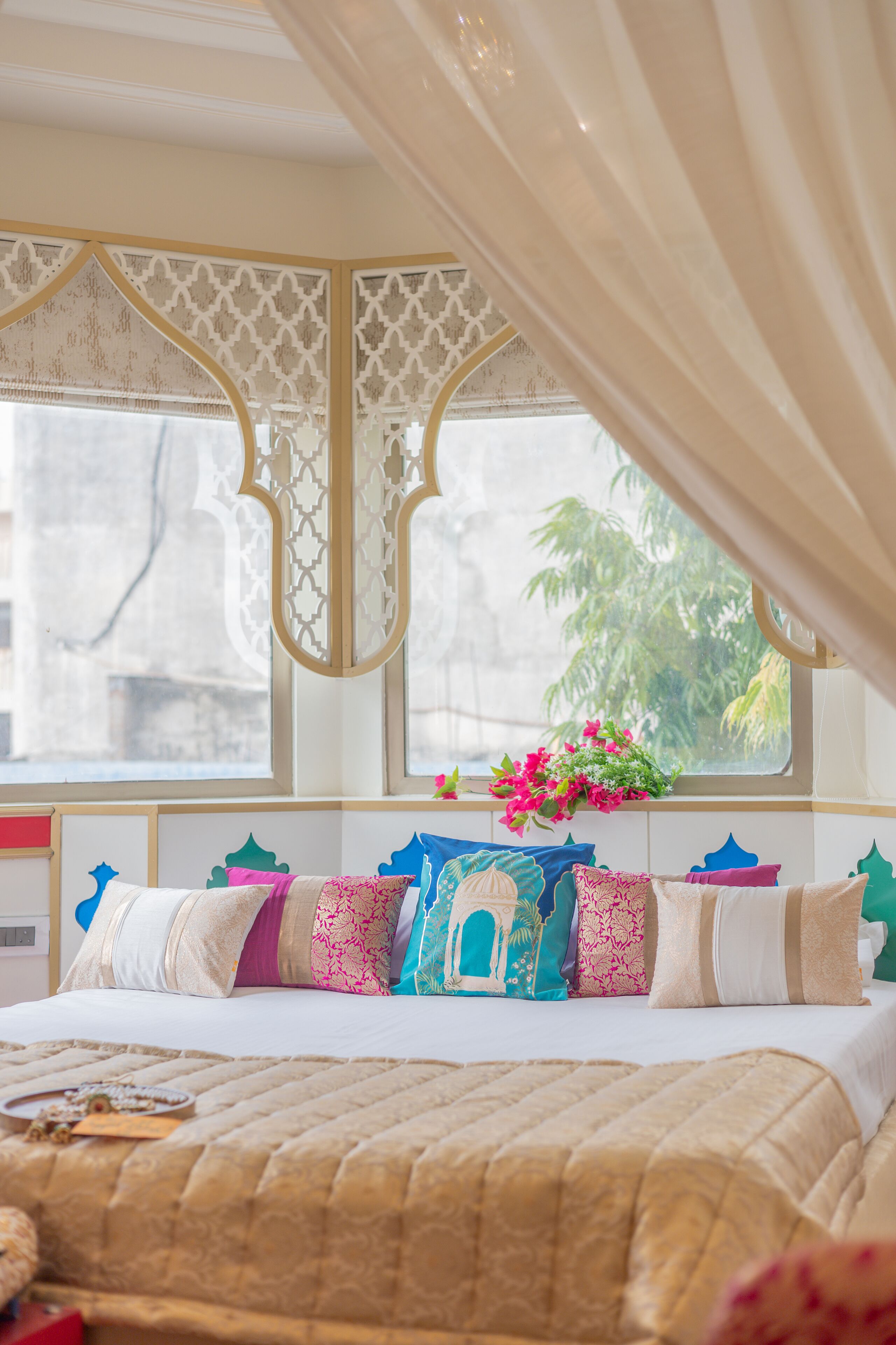 Una cama con colcha color arena, una ventana con vista a palmeras y una cortina decorativa.