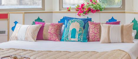 Una cama con colcha color arena, una ventana con vista a palmeras y una cortina decorativa.