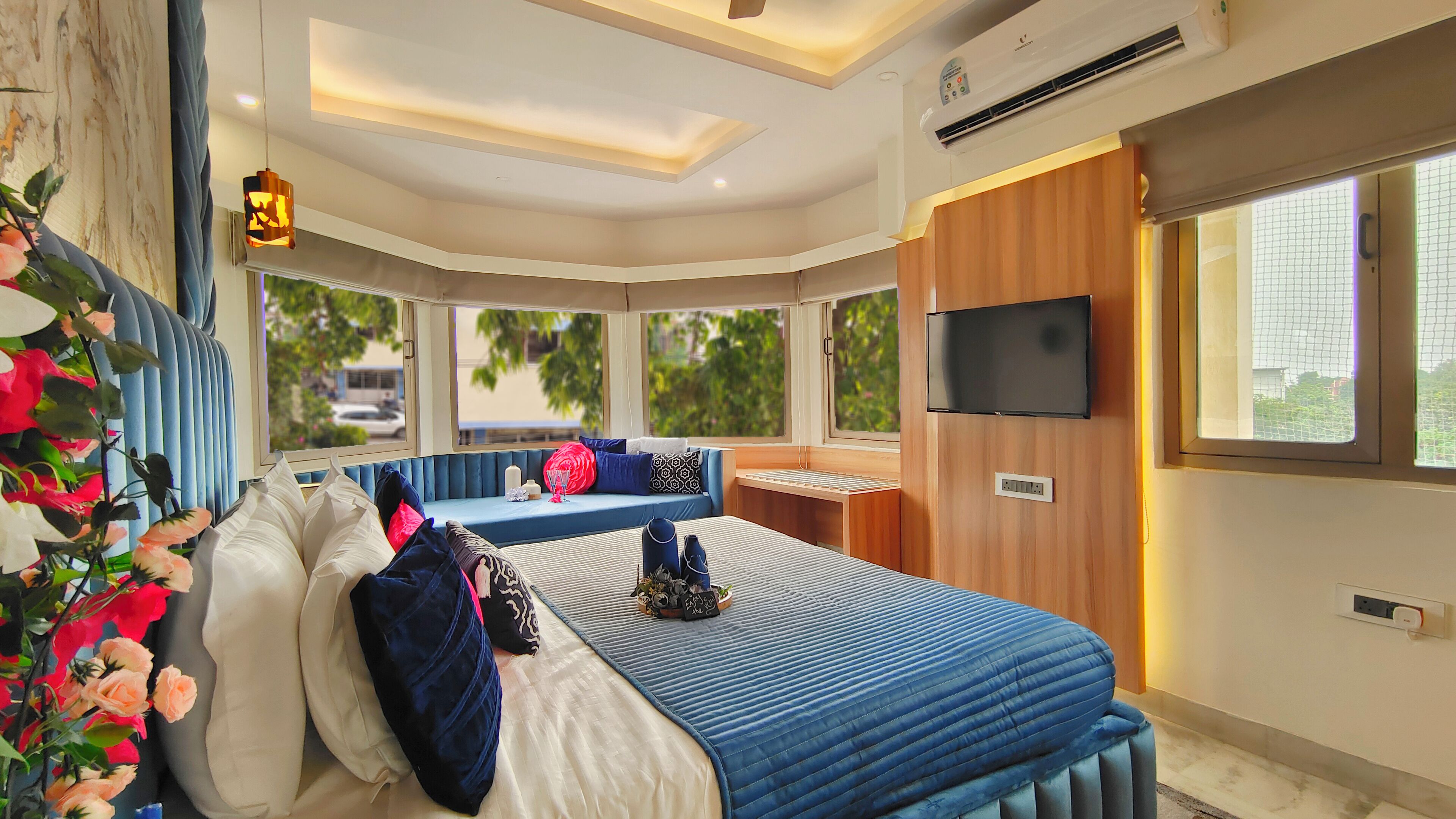 A modern bedroom with a large bed, a seating area, a TV, and a window with a view of greenery.