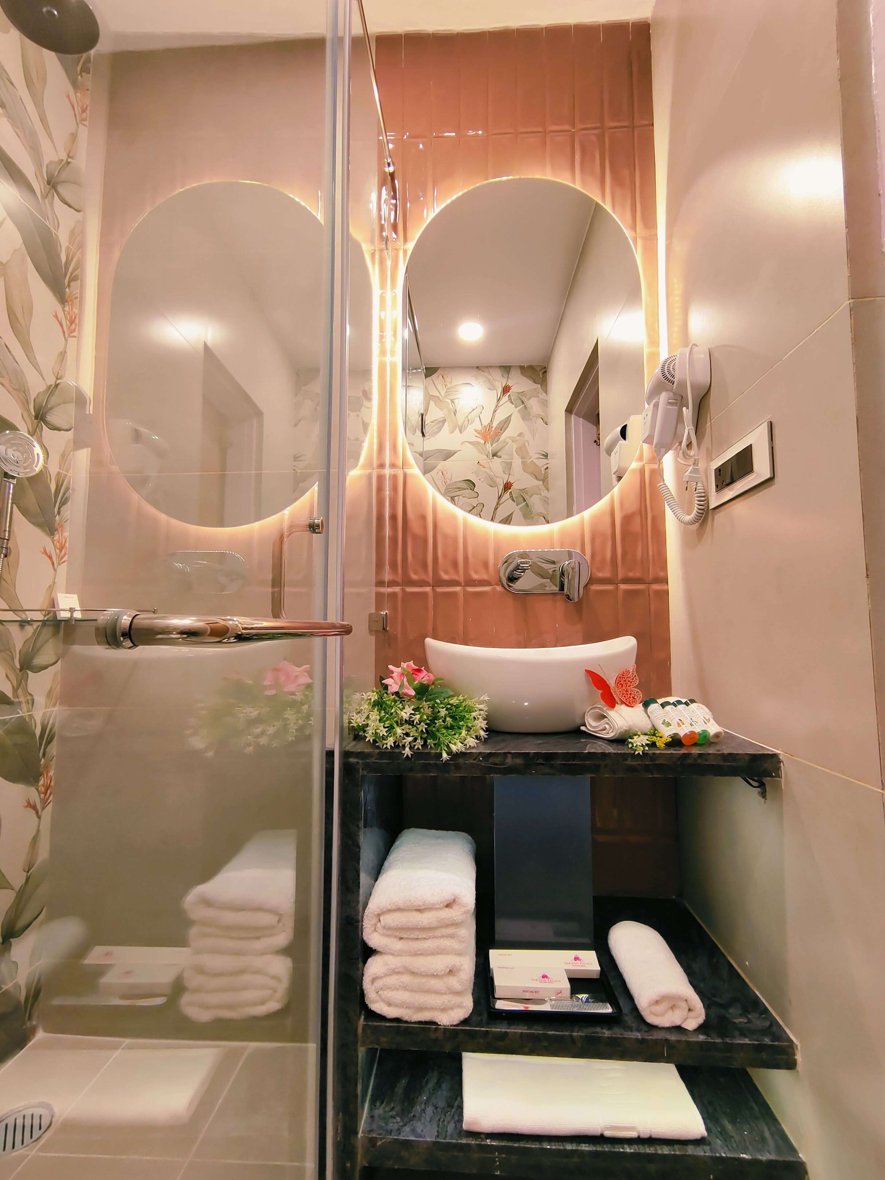 A modern bathroom with a glass shower enclosure, a large mirror, a sink with a decorative vase, and neatly folded towels on a shelf.
