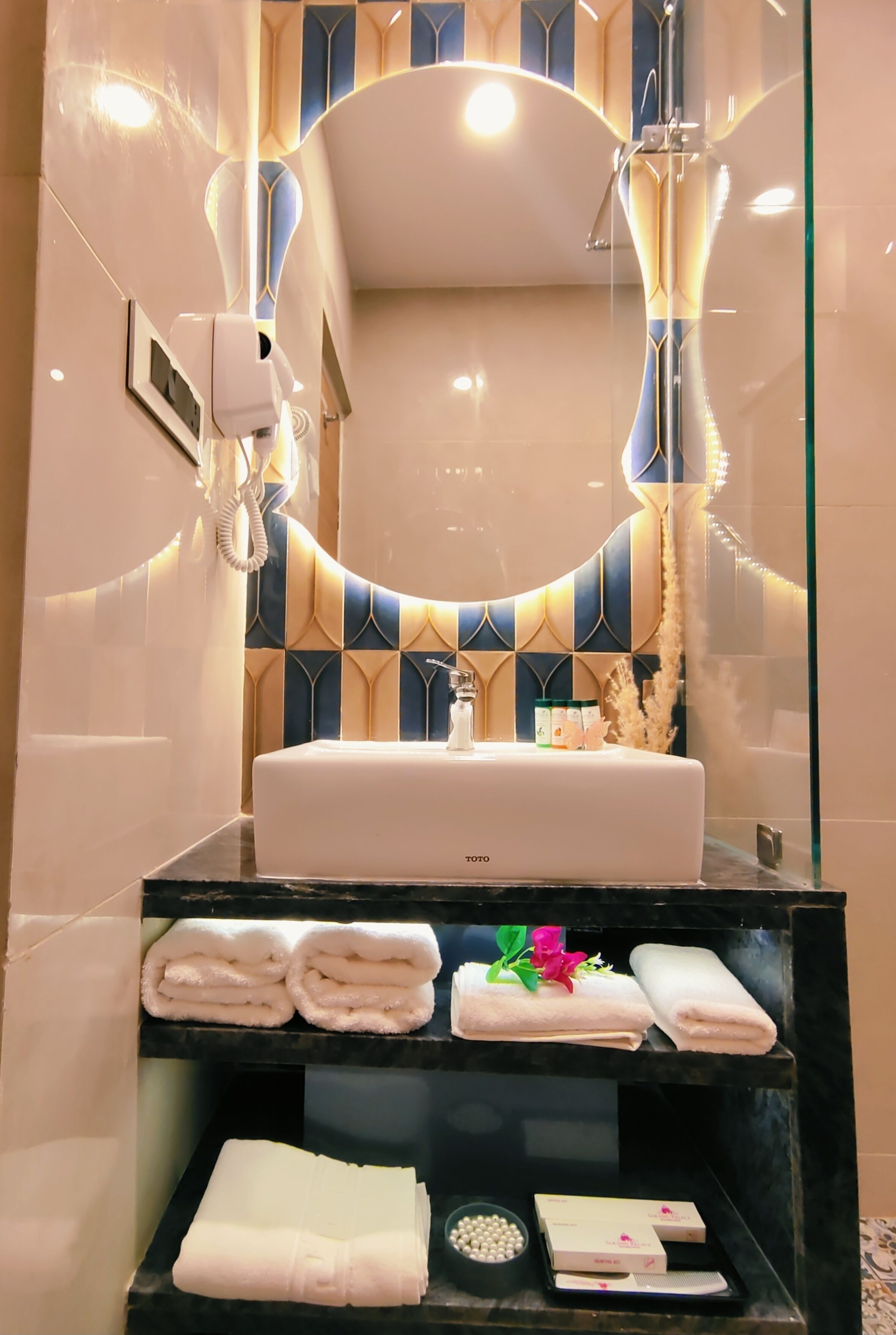 A modern bathroom with a large mirror, a white sink, and a black countertop. There are towels and toiletries on the counter.
