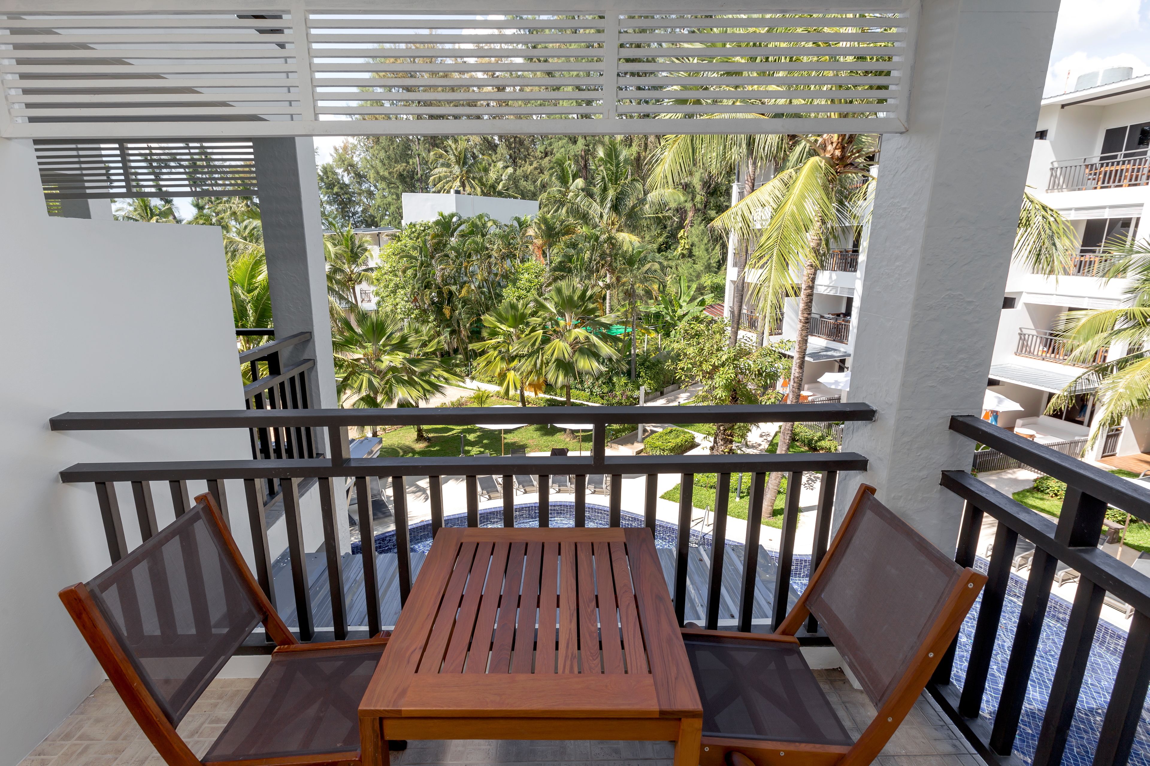 A balcony with a wooden table and chairs, overlooking a pool and lush greenery.