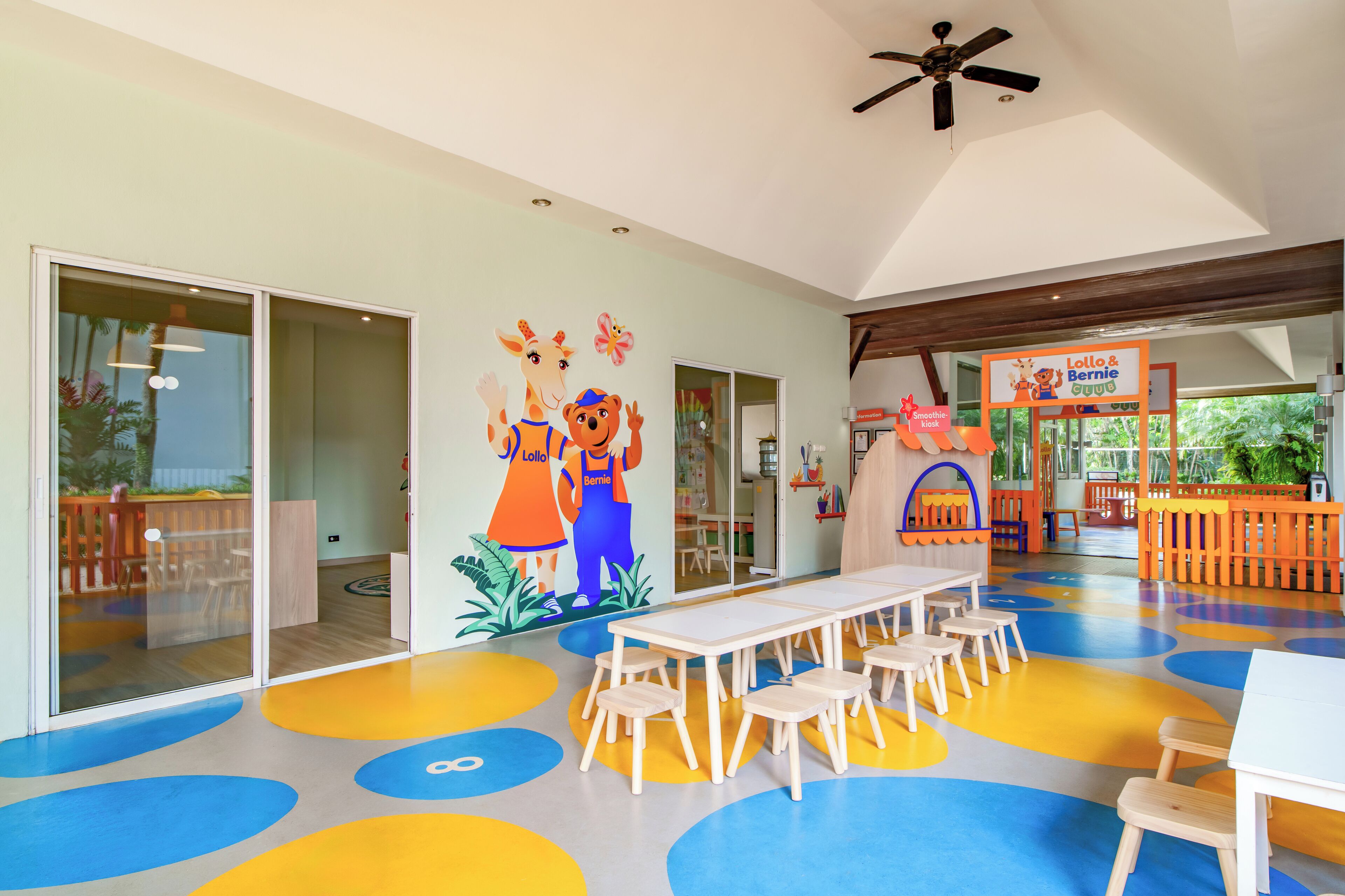Un área de juegos infantiles con piso colorido, un mural de Lollo y Bernie, y una mesa con sillas.