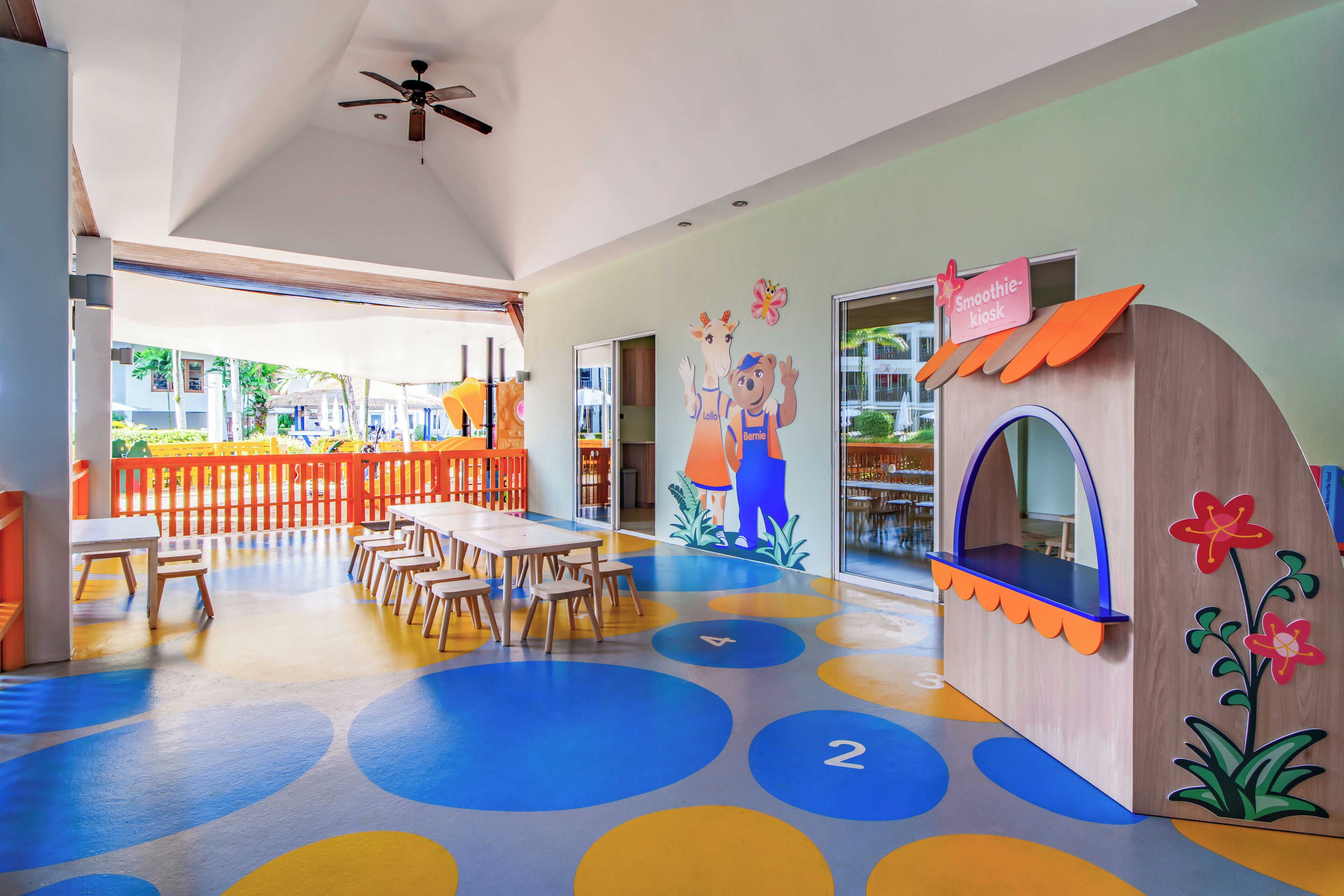 Un espace de jeux pour enfants avec un sol coloré, des tables, des chaises et une maison de jeu.