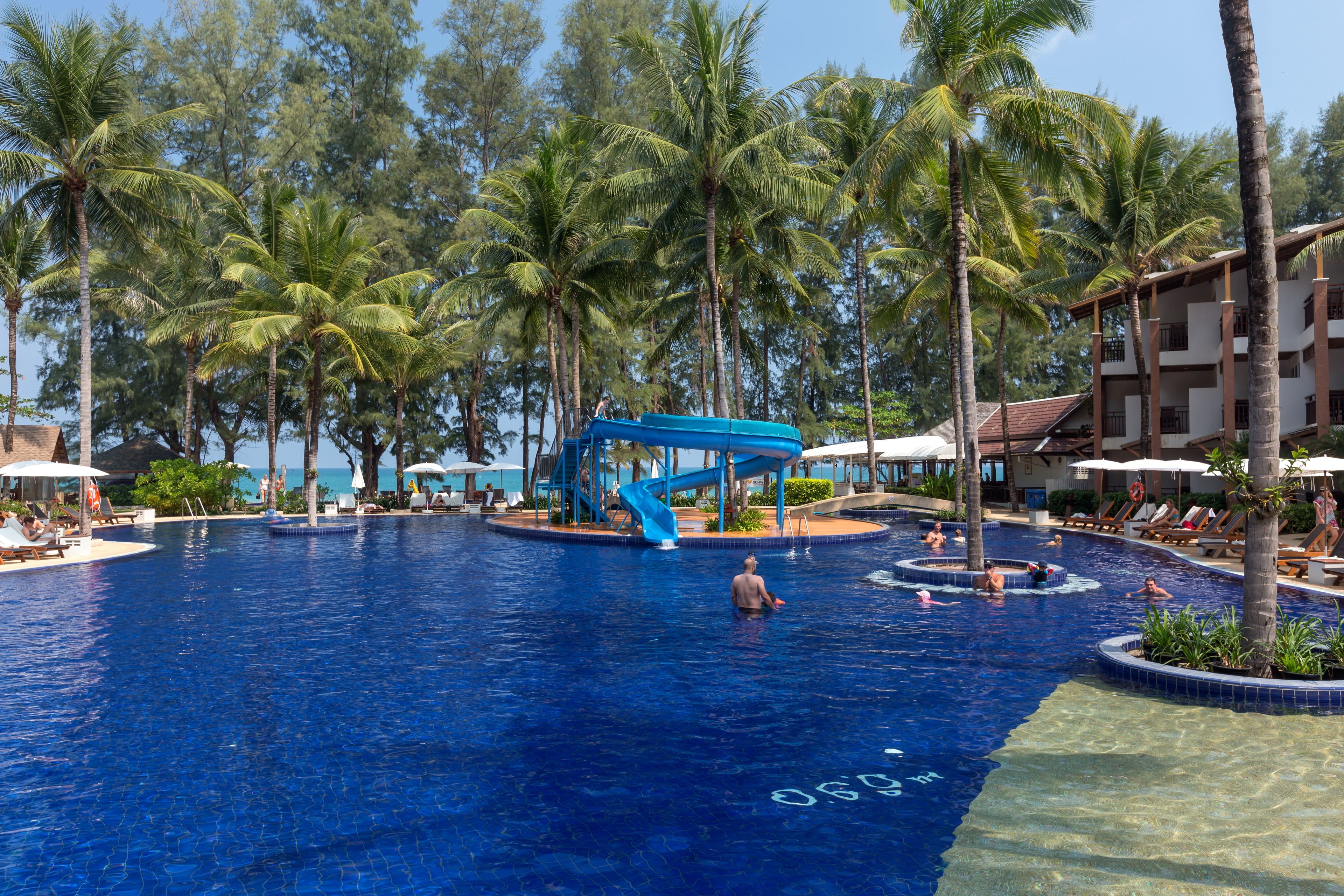 A large swimming pool with a blue slide, surrounded by palm trees and lounge chairs.