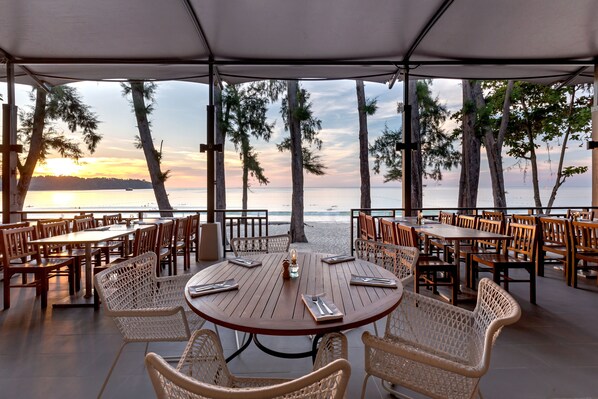 An outdoor dining area with round tables and wicker chairs, overlooking a beach and ocean at sunset. - Sunwing Bangtao Beach (Choeng Thale)