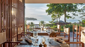 Una mesa de comedor de madera dispuesta con vasos, cubiertos y servilletas, con vistas a un paisaje marítimo y boscoso.