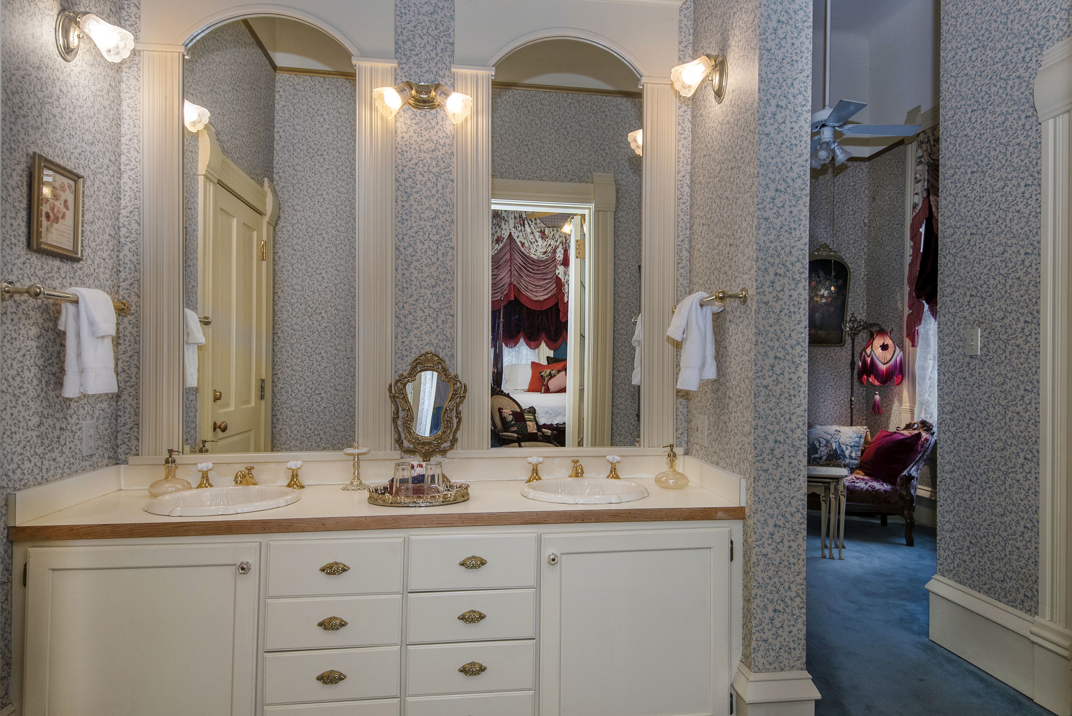 A bathroom with a large vanity, multiple mirrors, and a view into a bedroom with a bed and curtains.