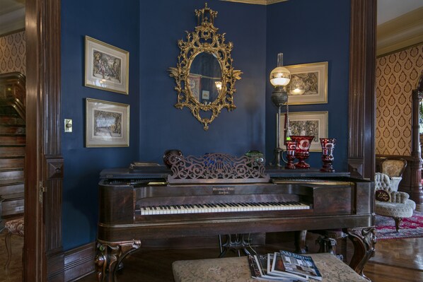 A grand piano in a room with dark blue walls, ornate gold-framed mirrors, and framed artwork.