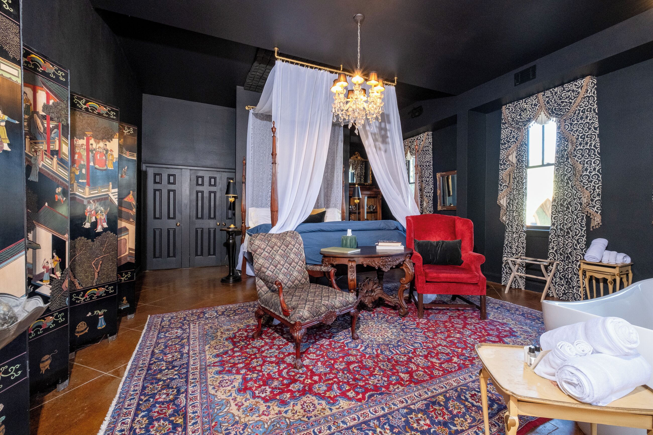 Una habitación con una gran silla ornamentada, un sillón rojo, una cama con dosel blanco, una araña y una alfombra estampada.