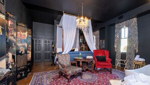 Ein Zimmer mit einem großen roten Sessel, einem blauen Sofa, einem hölzernen Stuhl, einem roten Stuhl, einem weißen Himmelbett, einer Chandelieber, einem gemusterten Teppich und einem kleinen Tisch mit Handtüchern.