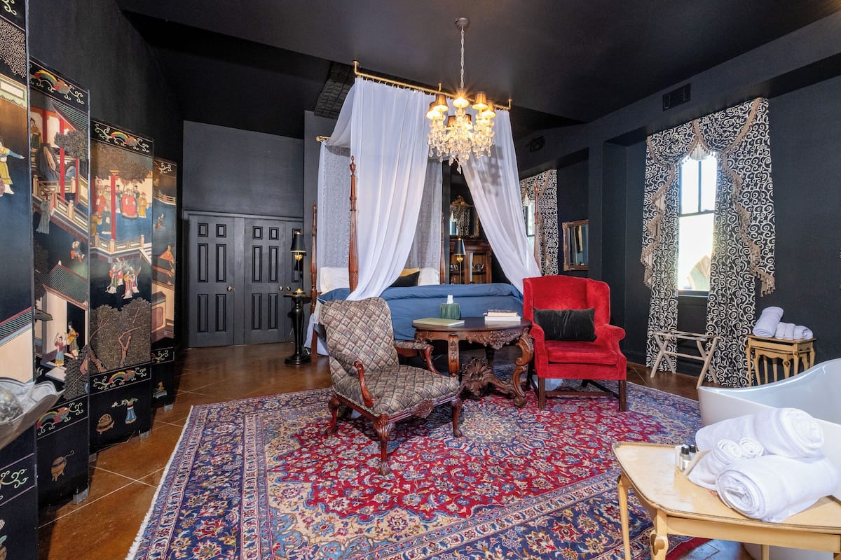 Une pièce comprenant un grand fauteuil rouge, un canapé bleu, une chaise en bois, une chaise rouge, un lit à baldaquin blanc, un lustre, un tapis à motifs et une petite table avec des serviettes.