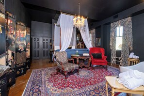 Een kamer met een grote, versierde stoel, een rode fauteuil, een bed met wit baldakijn, een kroonluchter en een kamerkleed met dessin.