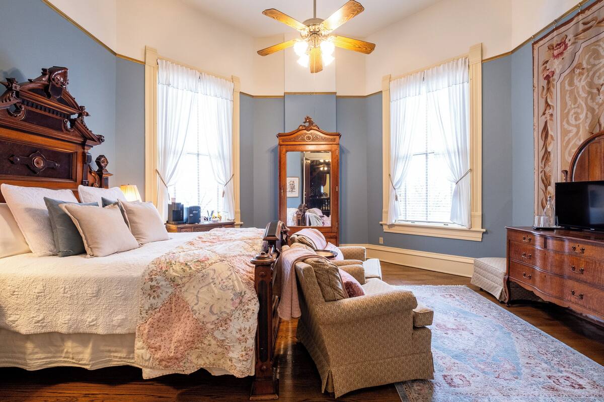 Une chambre à coucher avec un grand lit, une commode en bois, une chaise, un miroir et un ventilateur de plafond.