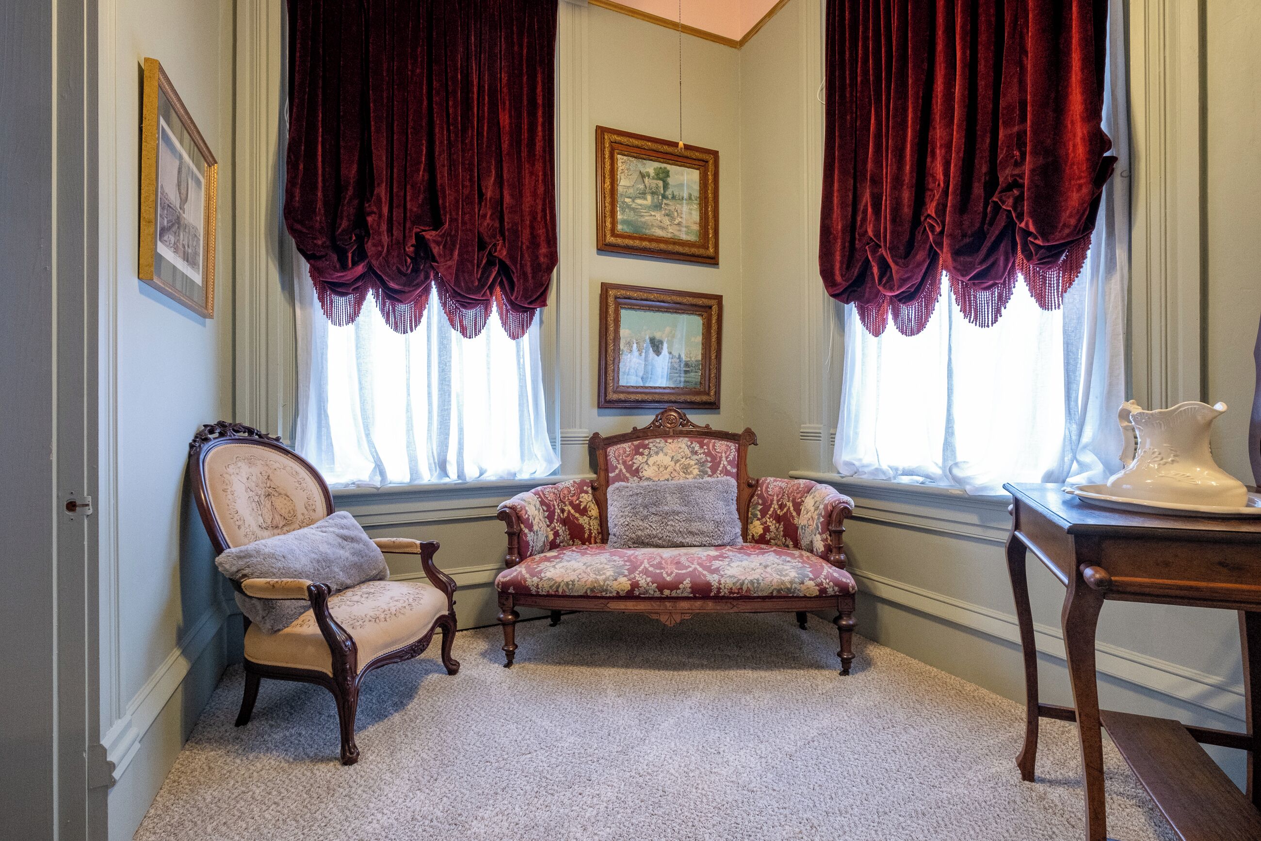 A room with two vintage chairs, a table with a pitcher, and two framed pictures on the wall.