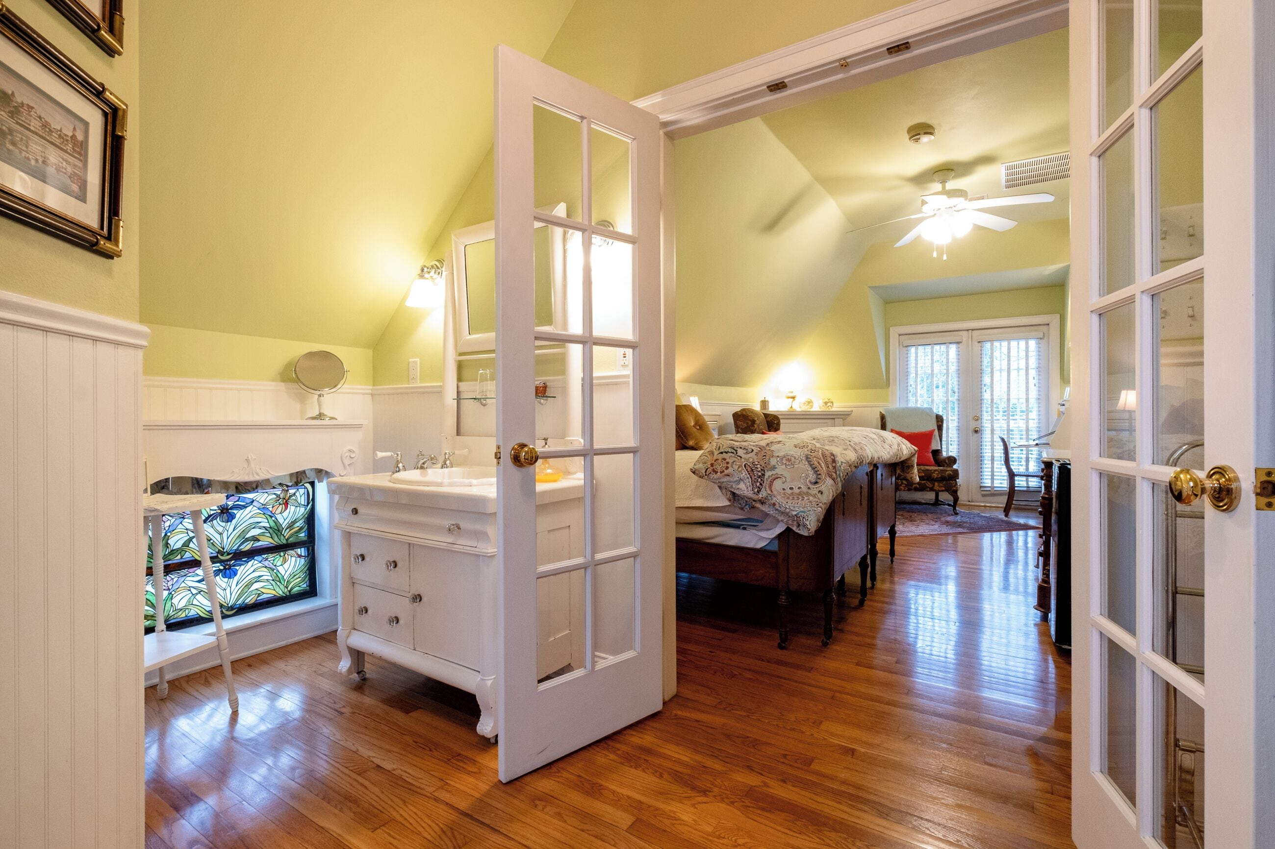 Una habitación con piso de madera, un tocador blanco con espejo, cama con colcha estampada y ventana con diseño de vitral.