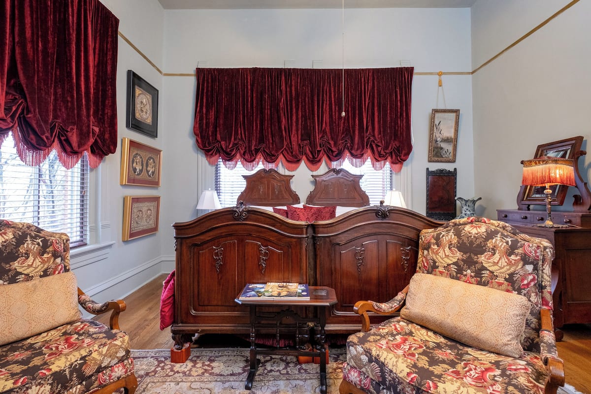 Une pièce comprenant un grand lit en bois, deux fauteuils rembourrés, une petite table et une lampe.