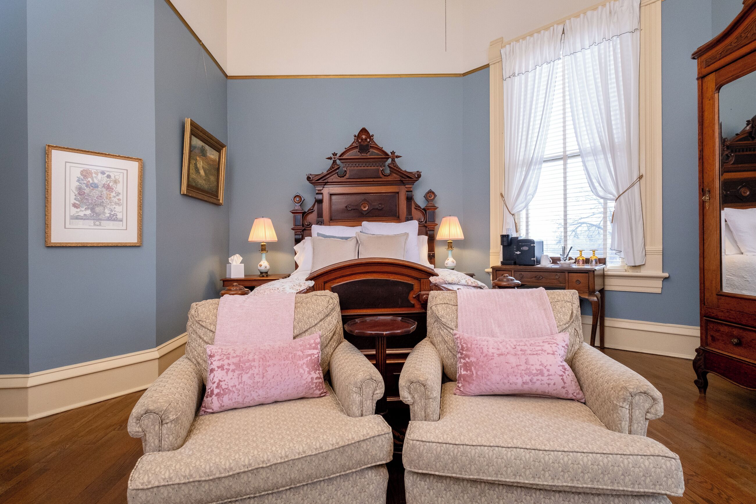 Ein Schlafzimmer mit einem großen Bett, zwei Sesseln, einem Schminktisch und einem Fenster mit Vorhängen.