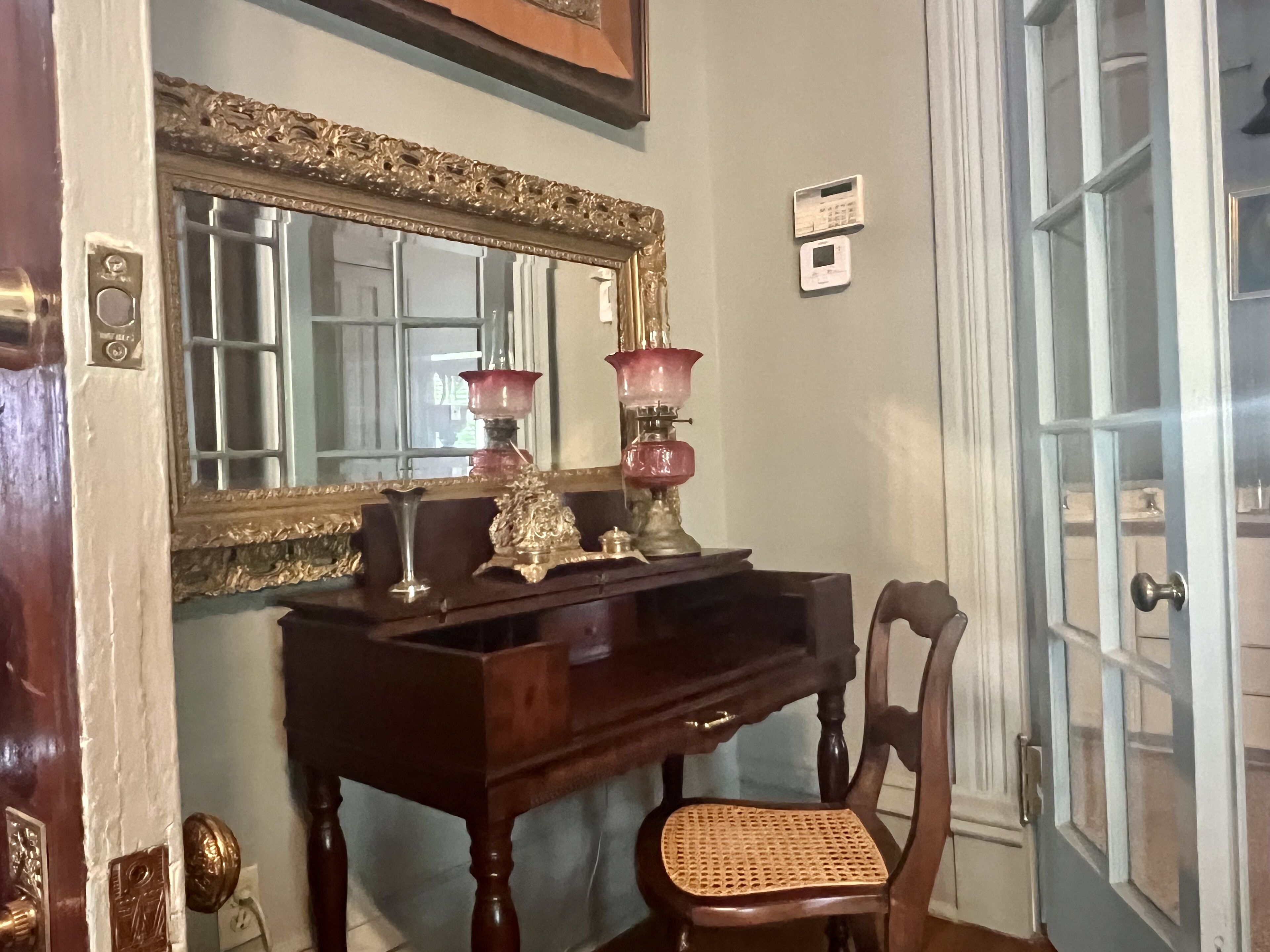 A room with a wooden desk, a chair, a mirror with an ornate frame, and a door with a glass panel.