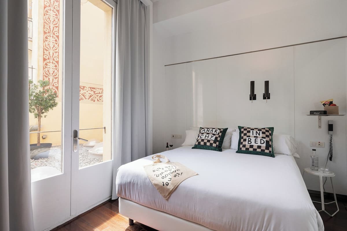 A modern bedroom with a large bed, two pillows, a bedside table with a phone, and a view of a balcony with a plant.