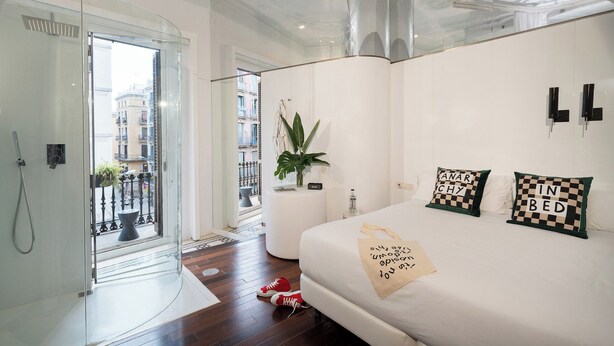 A modern bedroom with a glass shower, a white bed with checkered pillows, and a view of the city through a glass door. - Chic&Basic Born Boutique Hotel (Barcelona)