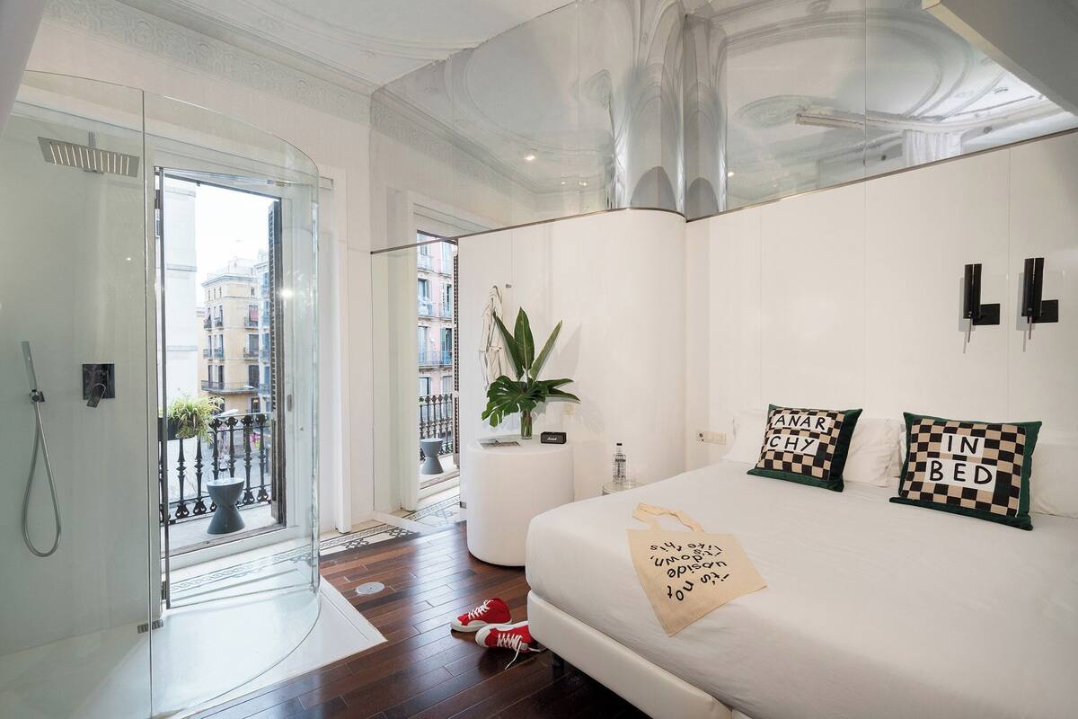 A modern bedroom with a glass shower, a white bed with checkered pillows, and a view of the city through a glass door.