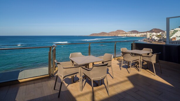 Una terraza con mobiliario exterior con vista al océano, cielo despejado y colinas lejanas.