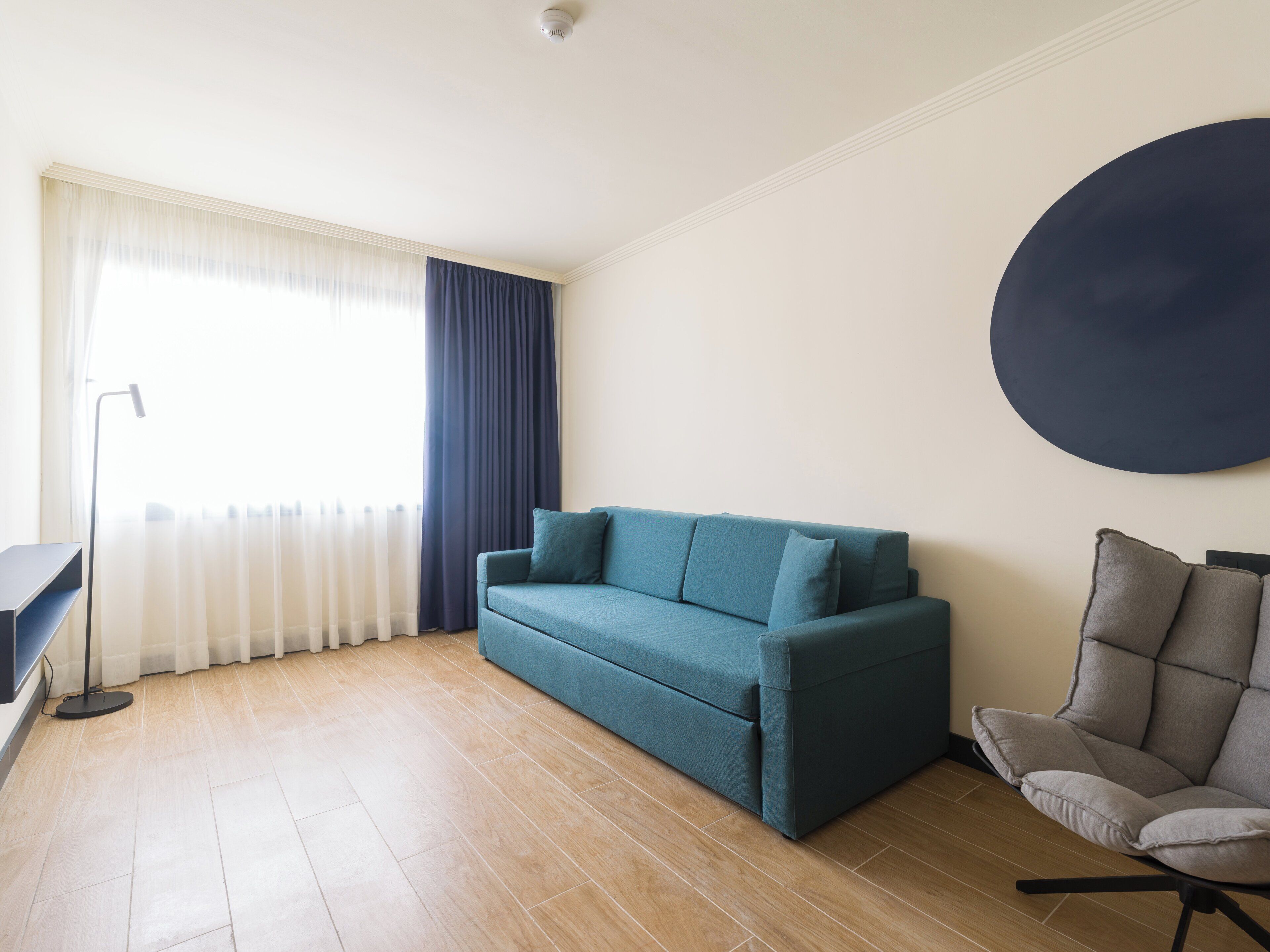 Uma sala de estar com sofá, poltrona, decoração circular na parede, luminária de chão e janela com cortinas.