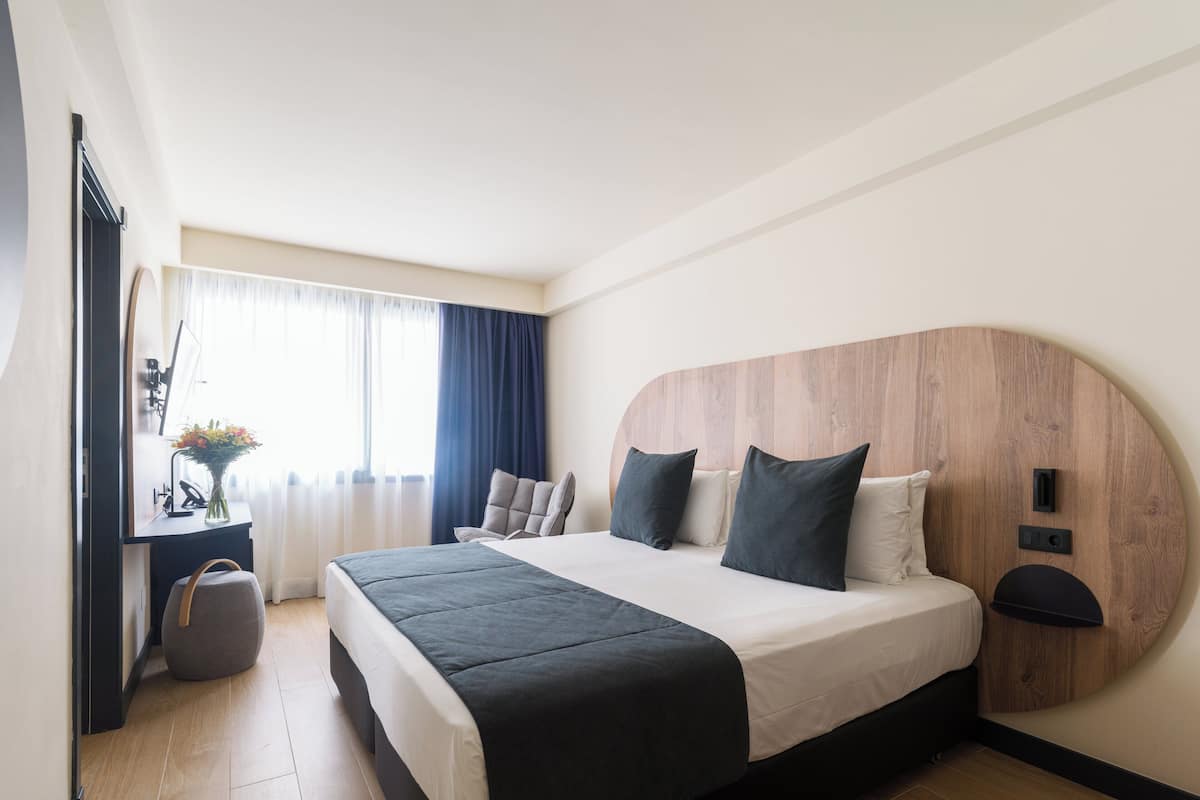 Ein modernes Hotelzimmer mit einem großen Bett, einem Schreibtisch mit Stuhl, einem kleinen runden Tisch und einem Fenster mit Vorhängen.