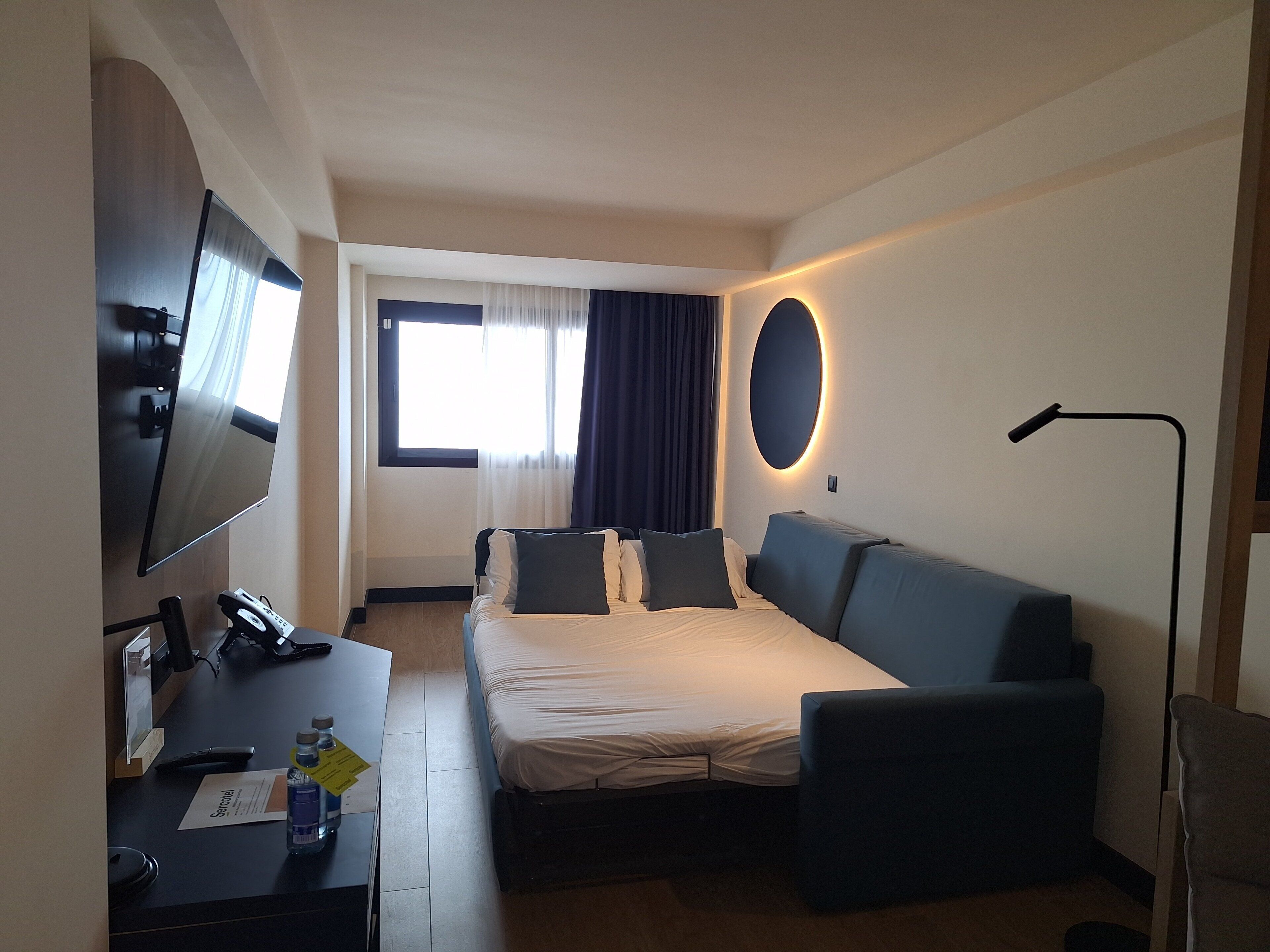 Habitación de hotel con cama, escritorio, computadora, televisor y ventana con cortinas.