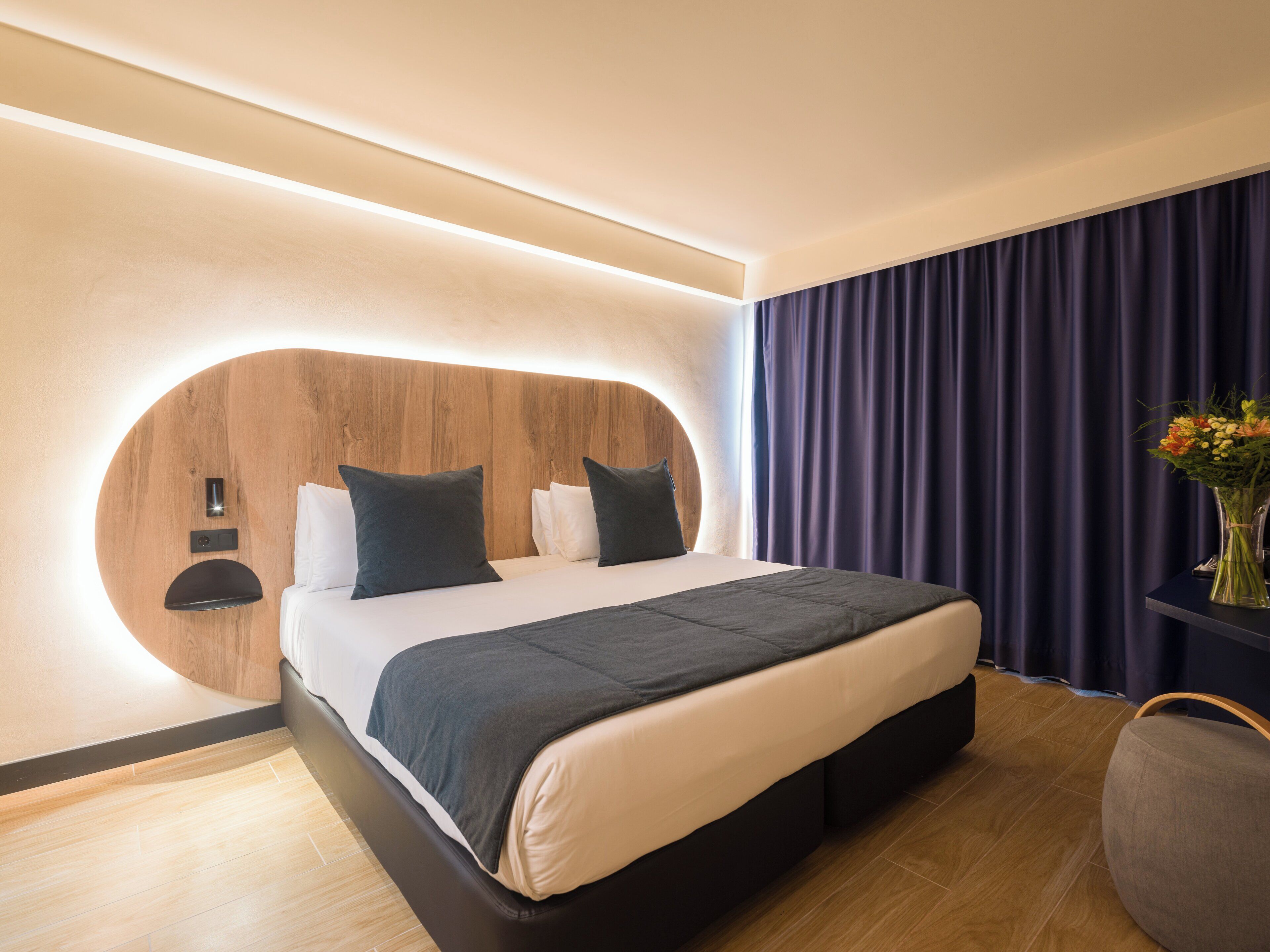 Moderni hotellihuone, jossa on kaareva sängynpääty, suuri sänky, yöpöytä ja maljakko, jossa on kukkia.