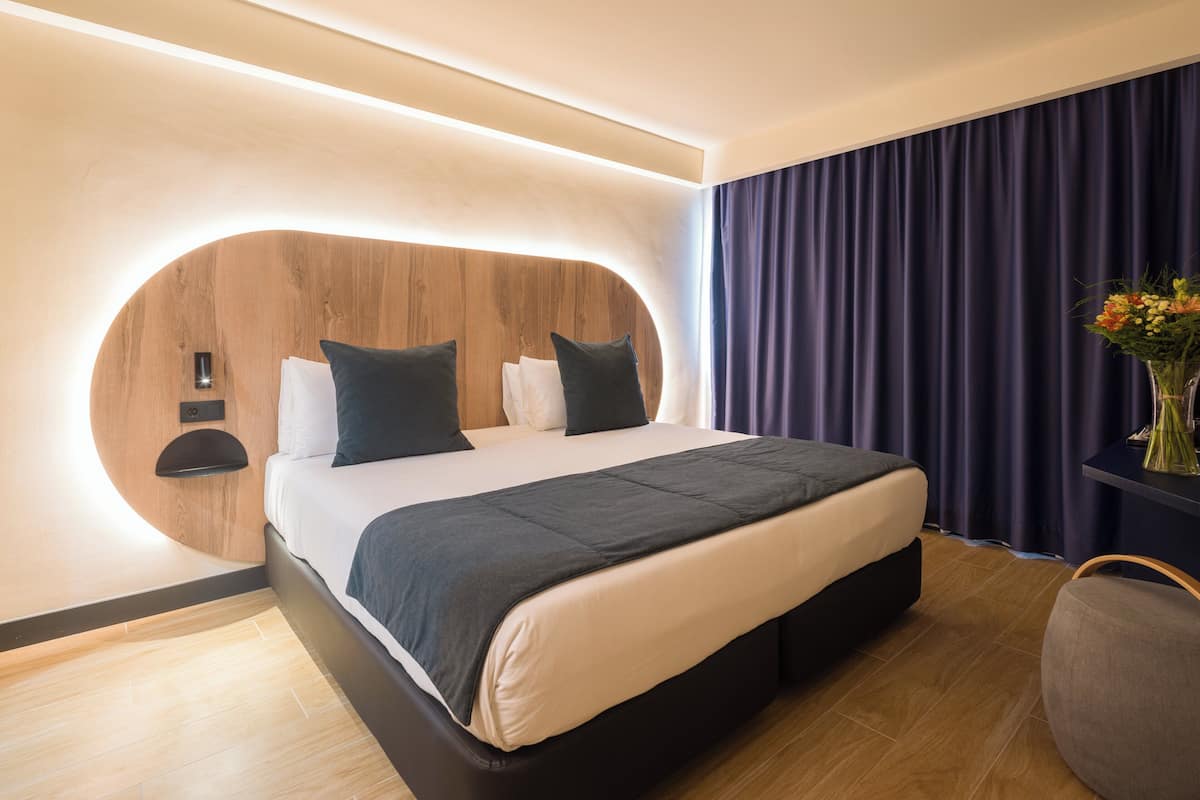 Ein modernes Hotelzimmer mit einem geschwungenen Kopfteil, einem großen Bett, einem Nachttisch und einer Vase mit Blumen.