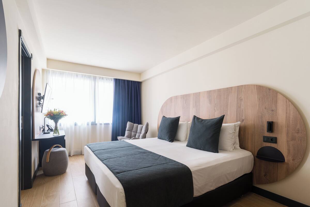 Una habitación de hotel moderna con una cama grande, un escritorio con una silla, una mesita redonda pequeña y un ventanal con cortinas.