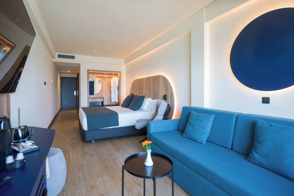 Una habitación de hotel moderna con una cama, un sofá azul, una pequeña mesa con un florero y un escritorio con un hervidor.