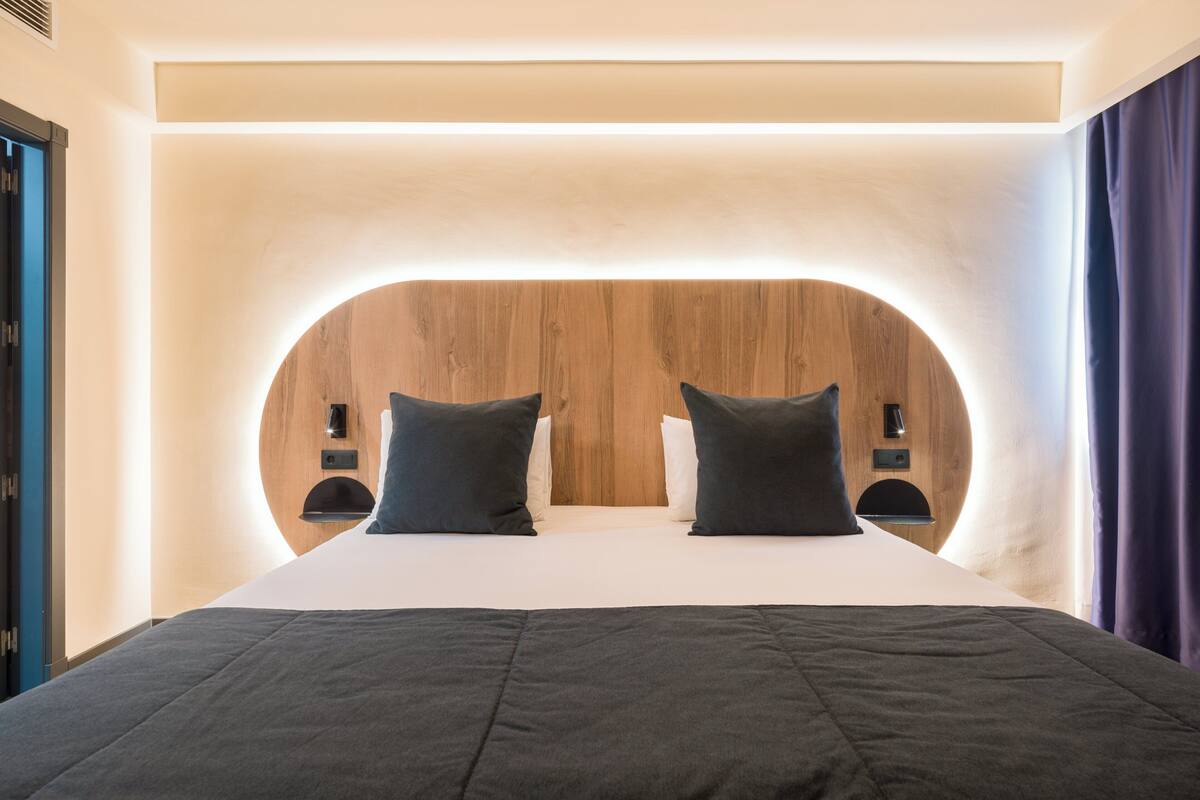 Una habitación de hotel moderna con una cabecera de madera curvada, dos mesitas de noche y una cama grande con ropa de cama oscura.