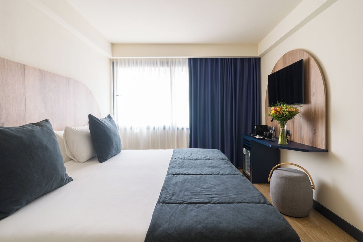 Una habitación de hotel moderna con una cama grande, un escritorio, un televisor de pantalla plana y una ventana con cortinas.