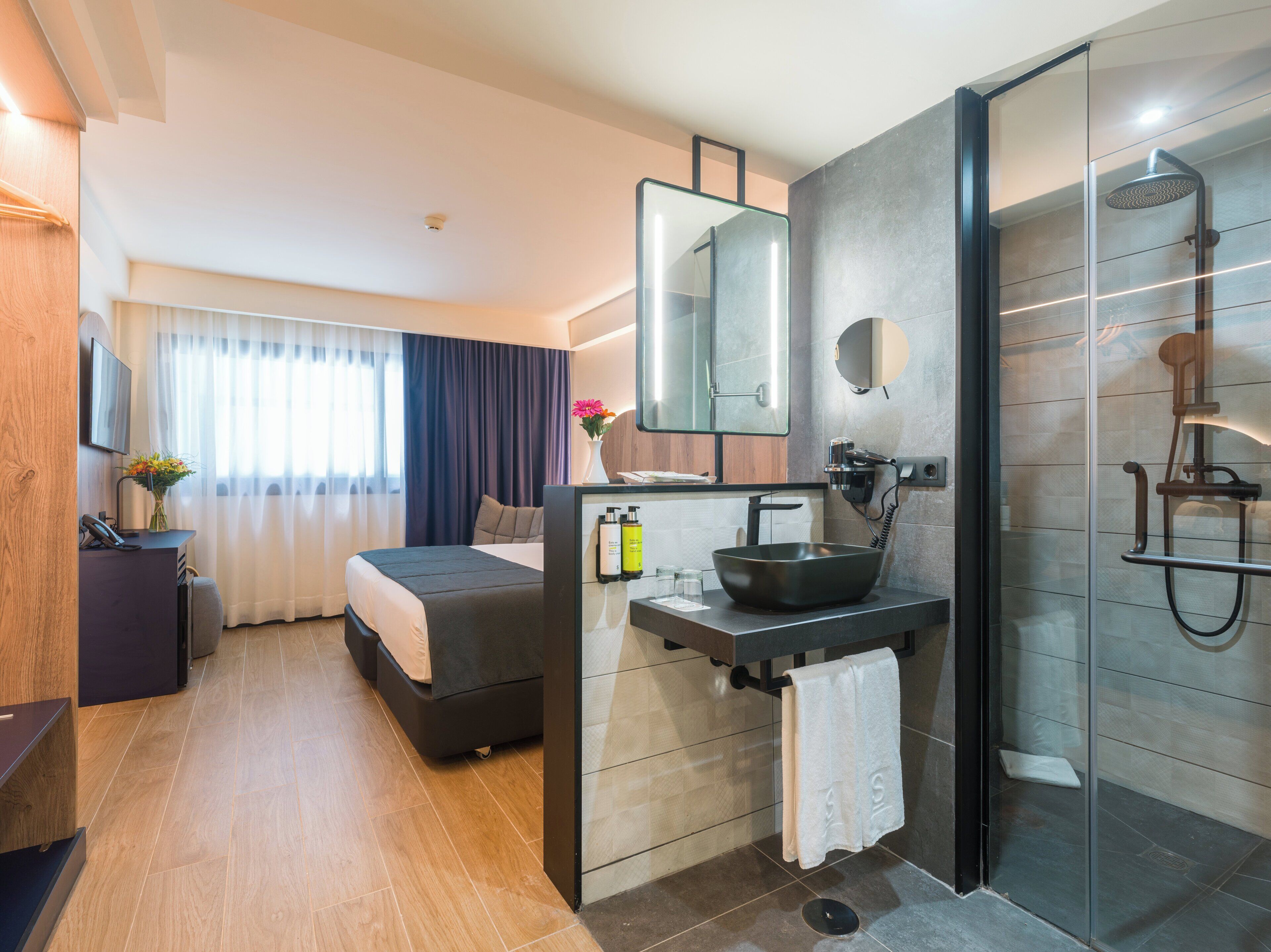 Una habitación de hotel moderna con cama, mesita de noche con teléfono, un escritorio pequeño con computadora, baño con lavamanos, espejo y ducha.