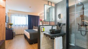 Una habitación de hotel moderna con cama, mesita de noche con teléfono, un escritorio pequeño con computadora, baño con lavamanos, espejo y ducha.