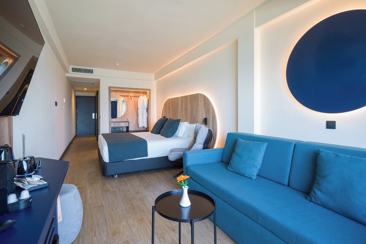 Ein modernes Hotelzimmer mit einem Bett, einem blauen Sofa, einem kleinen Tisch mit einer Vase und einem Schreibtisch mit einem Wasserkocher.
