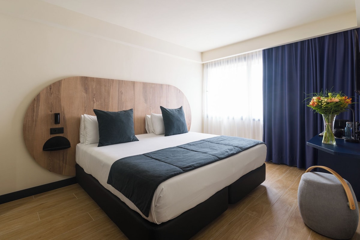 A modern hotel room with a wooden headboard, a large bed with a dark blanket, a bedside table with a vase of flowers, and a luggage storage unit.