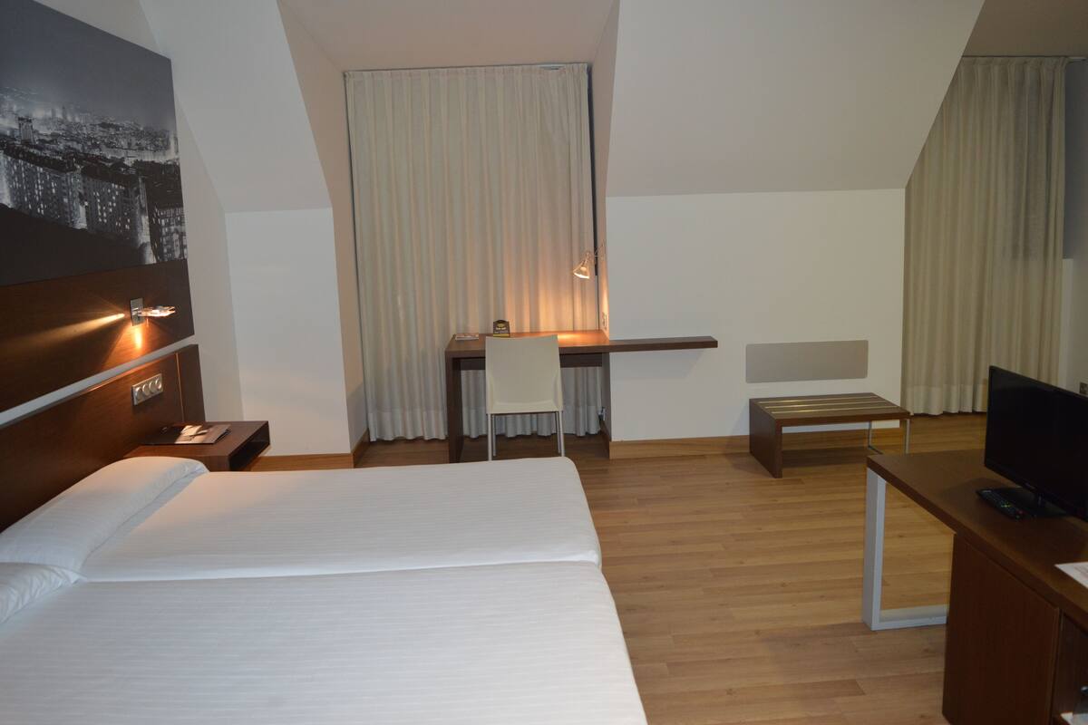 Una habitación de hotel con una cama grande, un escritorio con silla, televisión y ventana con cortinas.