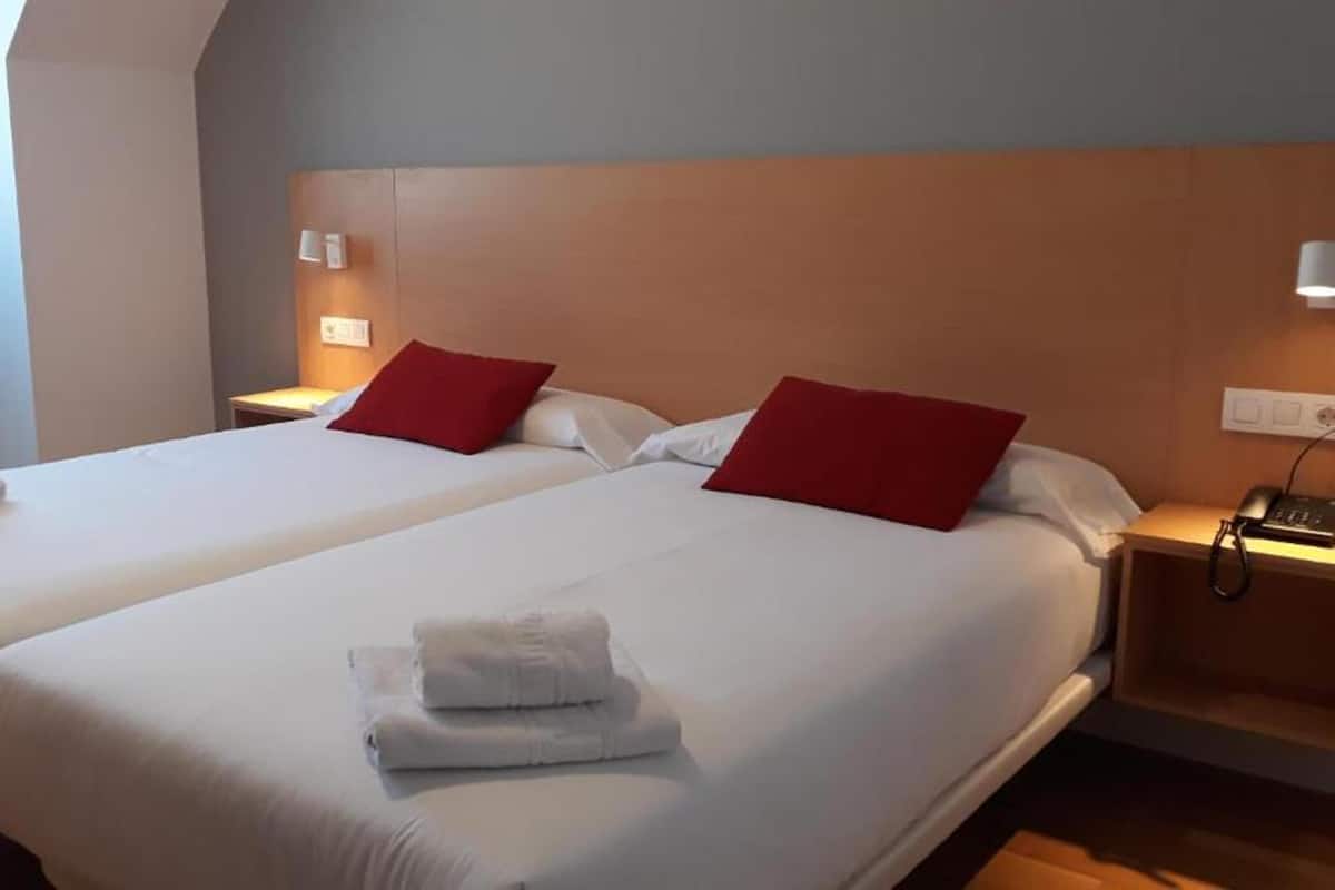 Habitación de hotel con dos camas, cada una con ropa blanca y almohadas rojas. Hay toallas sobre las camas y lámparas de noche a la vista.