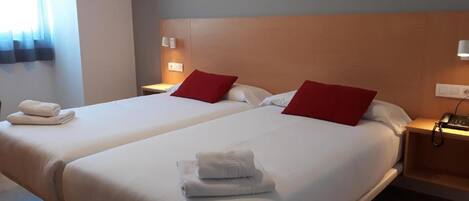 Habitación de hotel con dos camas, cada una con ropa blanca y almohadas rojas. Hay toallas sobre las camas y lámparas de noche a la vista.