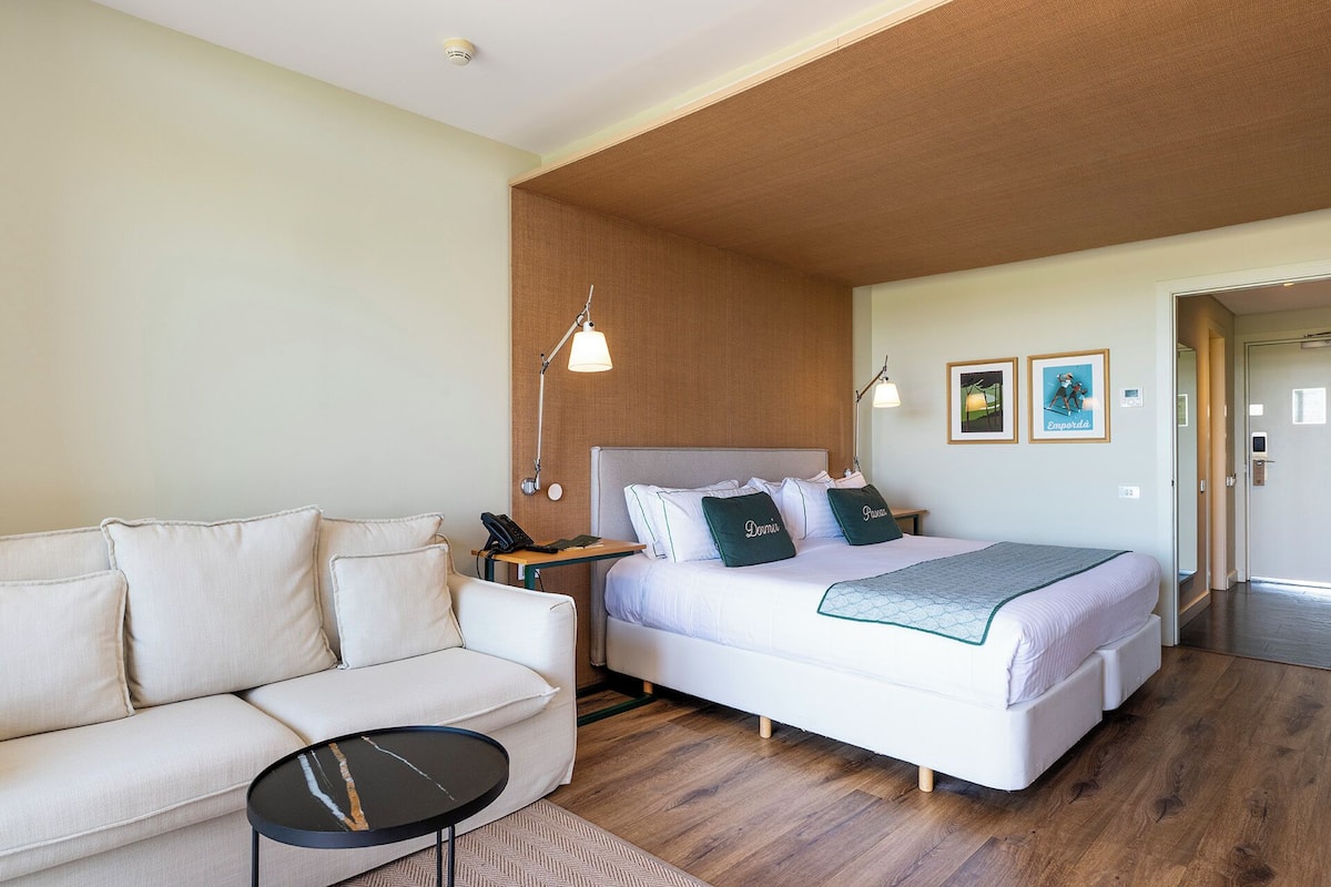 A bedroom with a bed, a sofa, a bedside table, a lamp, and two framed pictures on the wall.