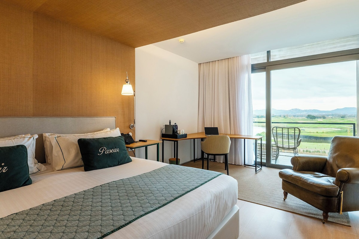 A hotel room with a bed, desk, chair, and a view of the countryside.